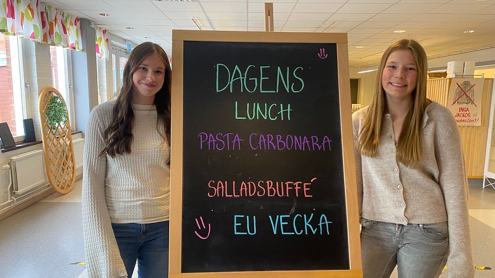 Två personer står vid en meny-tavla där det står ”Dagens lunch: Pasta carbonara, salladsbuffé, EU-vecka”.