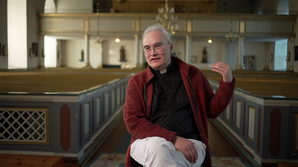 Prästen Oliver Li i Forsa kyrka med tomma bänkar och ljuskrona i bakgrunden.