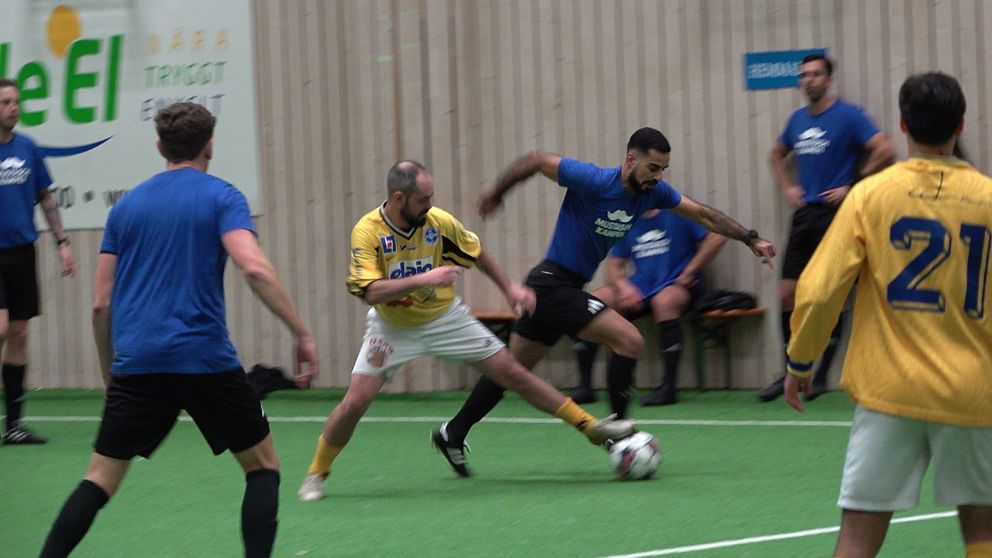 En inomhusfotbollsmatch med spelare i gula och blå tröjor som tävlar om bollen. En spelare i gul tröja försöker ta bollen ifrån en spelare i blå tröja, medan de andra spelarna observerar från sidan. Matchen äger rum på en grön konstgräsplan inomhus. Bakgrunden visar en vägg med sponsorskyltar.