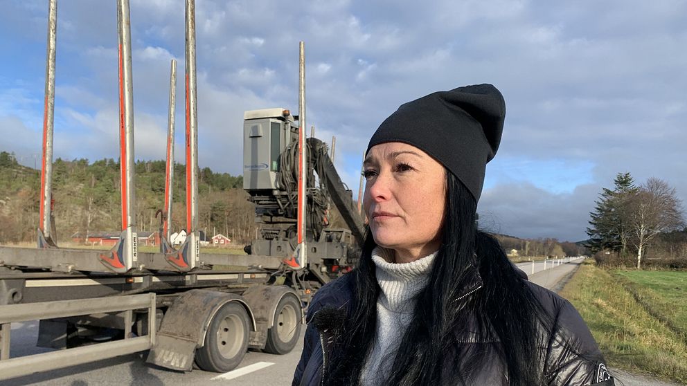 Malin Hansson står vid väg 650 där en timmerbil kör förbi.