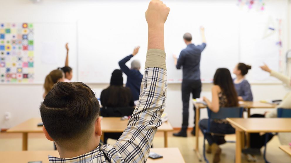 Elever som räcker upp handen. Lärare som står och skriver på whiteboardtavlan.