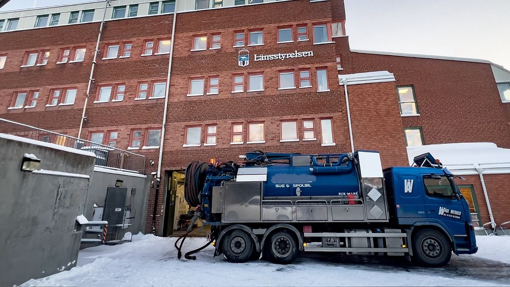 En snöklädd gård framför en tegelbyggnad med texten ”Länsstyrelsen” synlig. En pumpbil står utanför och pumpar vatten.