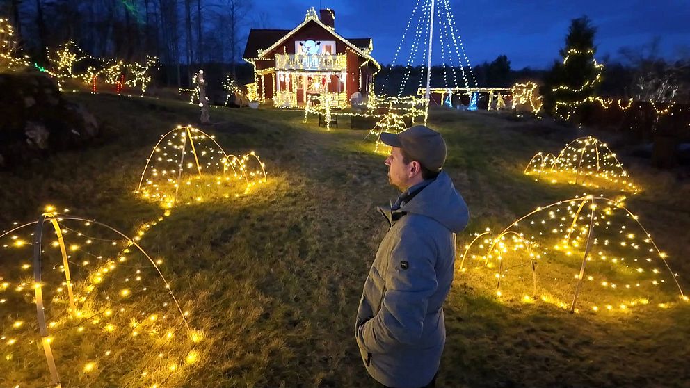 Kristoffer Rudin står på en grön gräsmatta som är täckt av strålande julbelysning. I bakgrunden syns ett rött hus dekorerat med flera ljus, och trädgården är prydd med lysande former i olika konfigurationer, inklusive bågar och spiraler. Stämningen är festlig och mysig, med en mörkblå himmel som ger en vacker kontrast till det gyllene ljuset.