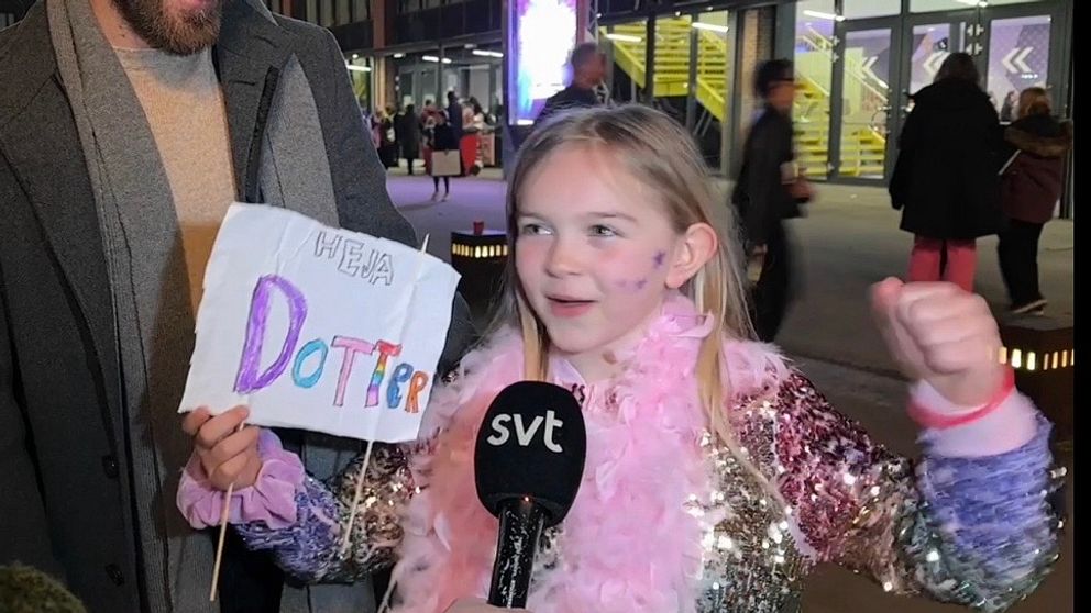 Tjej står med skylt och ser glad ut, håller upp armarna