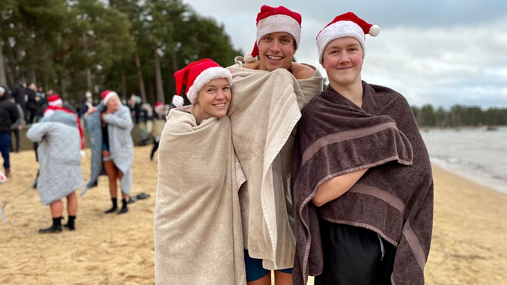 Tre personer iklädda tomteluvor står på en strand, insvepta i handdukar efter ett vintrigt bad. I bakgrunden syns andra deltagare som också deltar i aktiviteten. Stämningen är festlig med en tydlig julkänsla trots det kalla vädret.