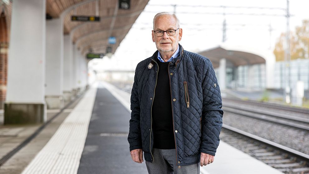 En person står på en tom tågplattform klädd i en mörk jacka och grå byxor. Plattformen är delvis skyddad av ett tak, och tågspåren sträcker sig bort i bakgrunden.