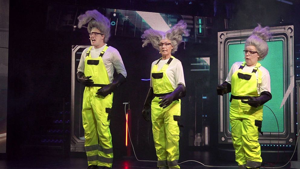 Tre personer i neonfärgade arbetskläder med reflexdetaljer står på en scen. De har alla liknande frisyrer i form av stora yviga gråa hår. Bakgrunden visar en teknologisk miljö med futuristiska inslag.