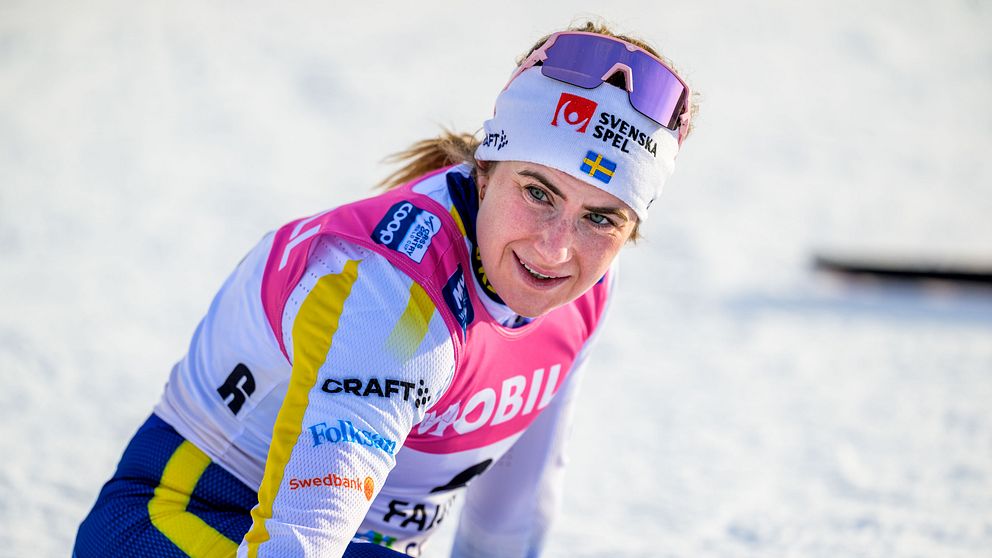 En tävlingsåkare i längdskidåkning utomhus, klädd i färgglad tävlingsdräkt med synliga sponsornamn, sitter på knä i snön.