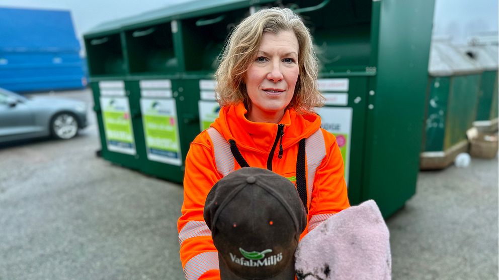 En person i en orange arbetsjacka håller en svart keps med en logotyp och en grå handduk, med en avfallscontainer i bakgrunden.