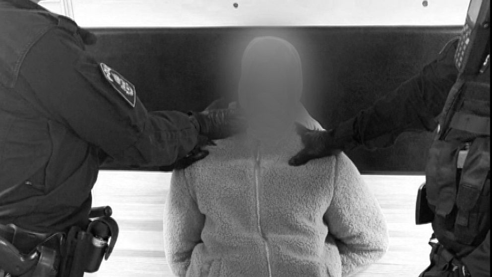 Den misstänkte mannen sitter bojad medan två poliser håller tag i honom. Mannens ansikte är pixlat och bilden svartvit.