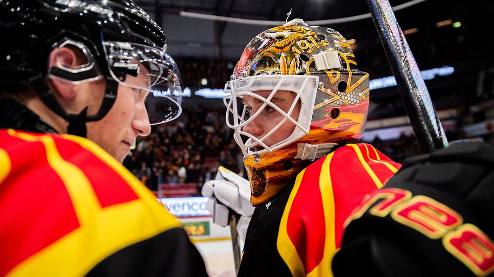 En grupp ishockeyspelare, med fokus på en målvakts hjälm som har en färgglad design med inslag av röd och gul. Spelarna bär matchtröjor i liknande färger, vilket skapar en enhetlig och dynamisk atmosfär. I bakgrunden syns en publik som är engagerad i matchen.