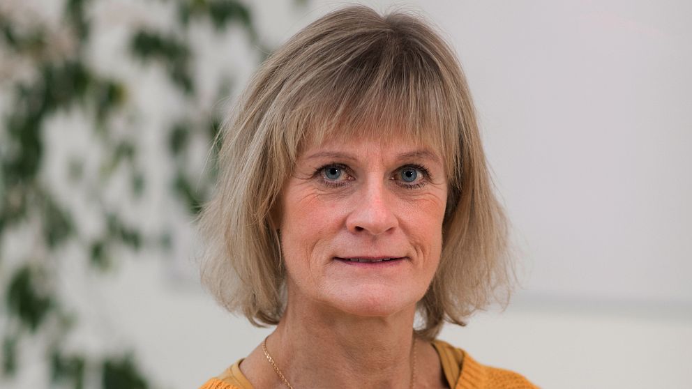 En person med blont hår klädd i en gul tröja, med en bakgrund av gröna växter och en vit vägg.