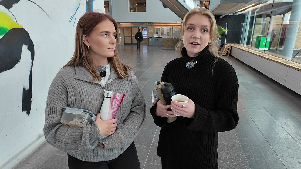 Två personer i en korridor, den ena håller en bok och den andra en kaffekopp.