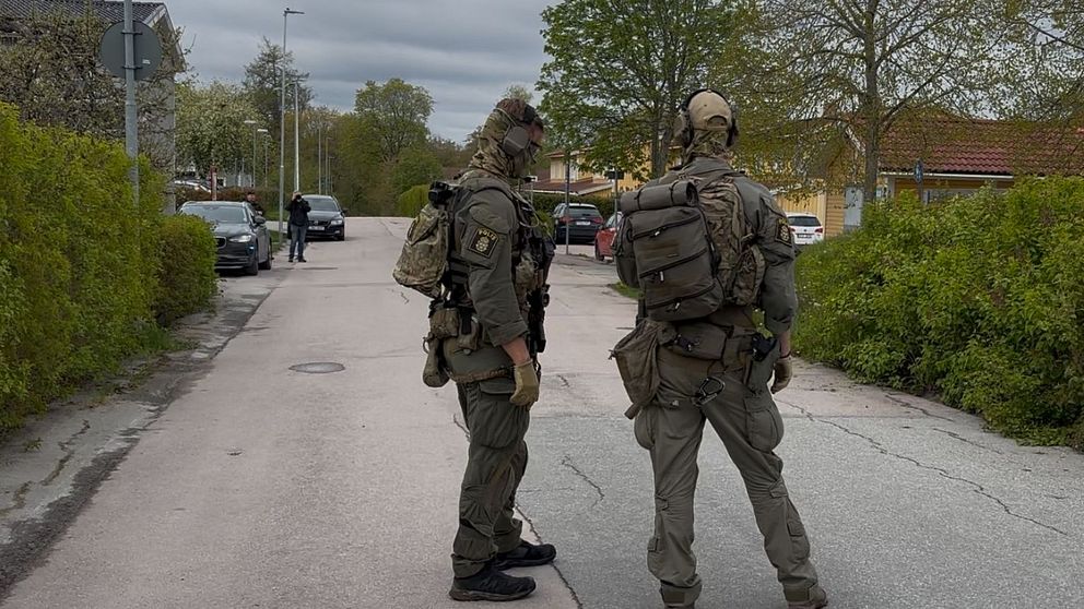 Två tungt beväpnade poliser står på en avspärrad gata i bostadsområde.