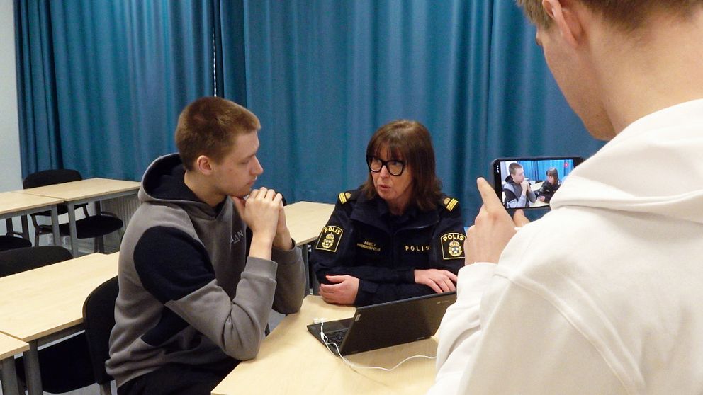 En polis sitter och pratar med en elev i ett klassrum. En annan elev filmar samtalet.