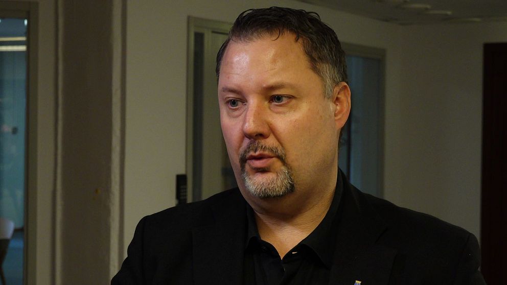 En man i mörk kostym står i en kontorsmiljö med glasväggar i bakgrunden. Han är delvis ur fokus, vilket skapar en mystisk känsla kring hans identitet. Det är en professionell och formell atmosfär i rummet.
