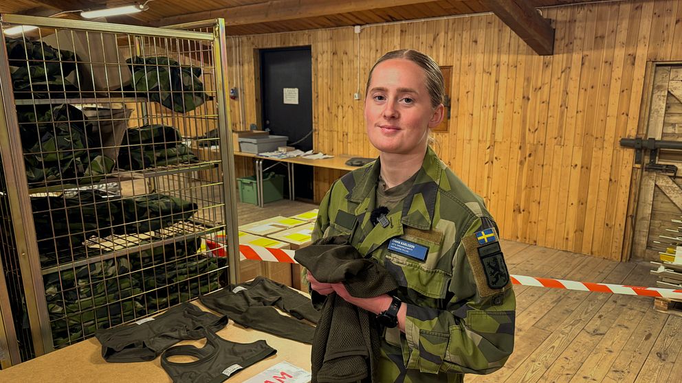 kvinna i gröna uniformskläder står inomhus och håller i en underställströja vid ett bord märkt ”DAM”