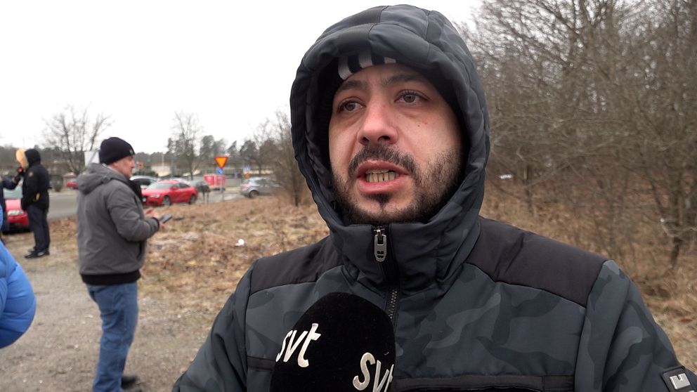 En person står utomhus i vinterkläder och håller en mikrofon med ”SVT” tryckt på, i förgrunden. I bakgrunden syns två personer och flera parkerade bilar längs en väg med träd utan löv.