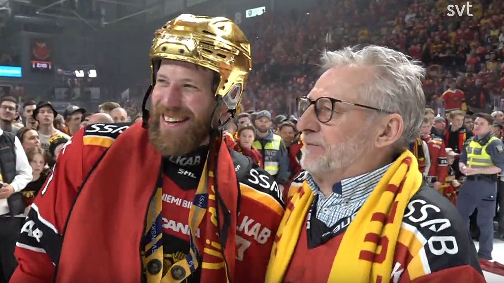 Två personer i hockeytröjor firar på isen efter match, omgivna av publik och funktionärer.