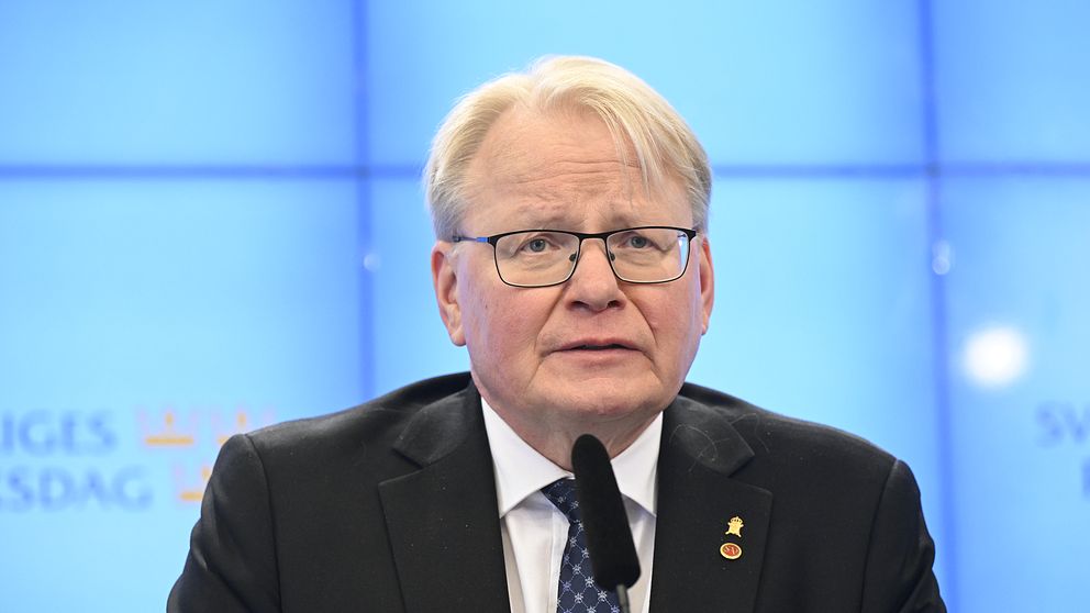 Peter Hultqvist talar i en mikrofon framför en blå bakgrund med Sveriges riksdags logotyp.