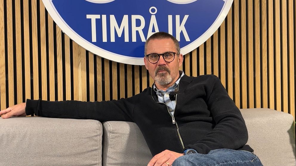 Person sitter avslappnat i soffa framför Timrå IK:s stora klubbmärke på väggen.