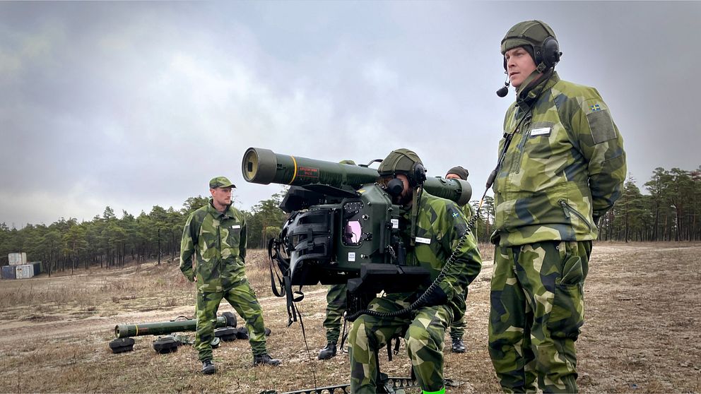 Soldater i militärkläder övar med en Robot 70 på ett öppet fält. En av soldaterna justerar en siktanordning medan de andra observerar och förbereder sig.