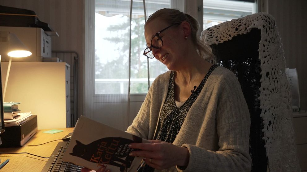 En person sitter vid ett skrivbord och läser en bok med en bild av en katt på omslaget. Skrivbordet är fyllt med böcker och kontorsmaterial.