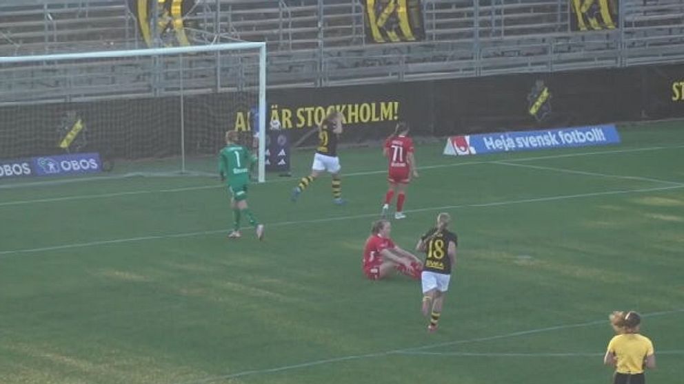 Fotbollsspelare på plan, en sitter ner nära målet. Domare i gul tröja i förgrunden.