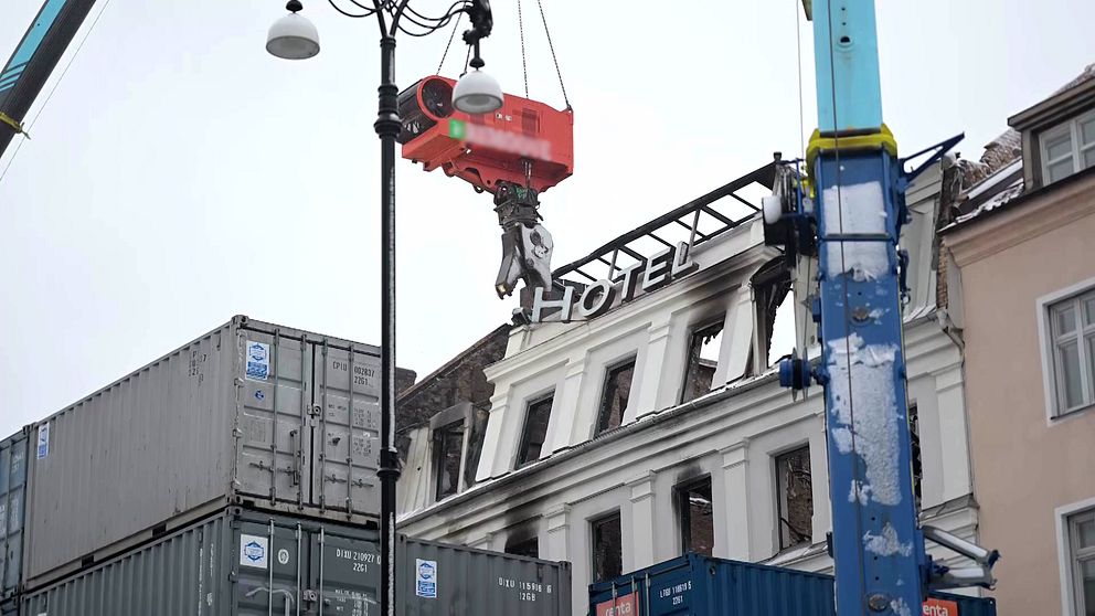 En kran som lyfter en röd kapsel över ett byggnadsområde, med en del av en byggnad i bakgrunden där skylten ”HOTEL” är synlig. Området är omgivet av fraktcontainrar.