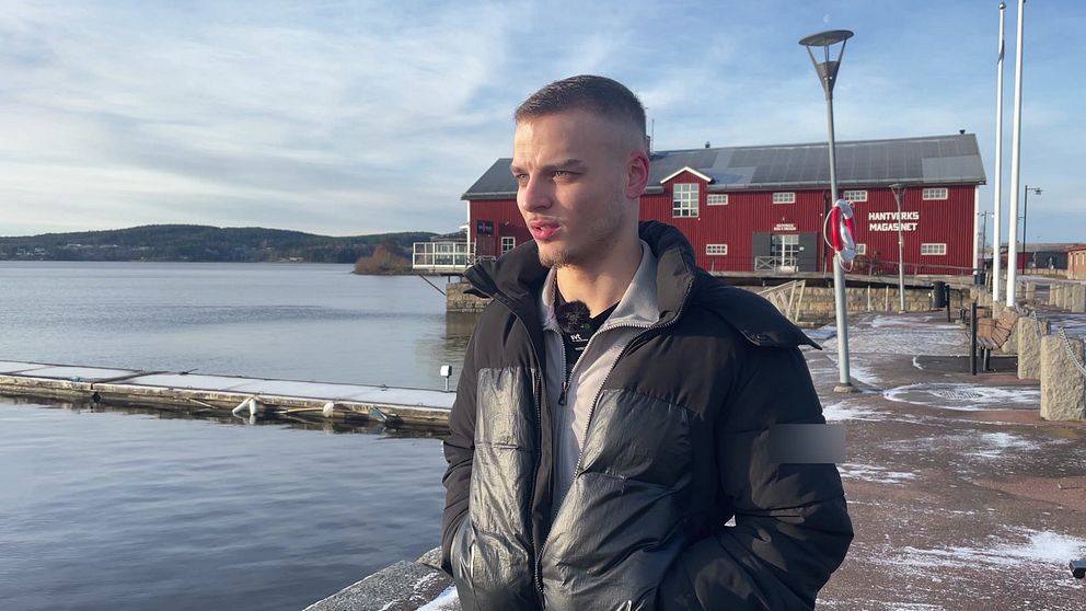 En person står på en brygga vid en sjö, klädd i en svart jacka. I bakgrunden syns ett rött hus och en lugn vattenyta med bergen i fjärran. Det är en klar dag med några moln på himlen.