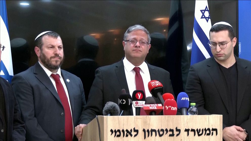 Tre personer med kippa står vid ett podium med mikrofoner och medieutrustning under en presskonferens. I bakgrunden syns en israelisk flagga och en skylt med hebreisk text, vilket indikerar att evenemanget äger rum i Israel. Personerna är formellt klädda i kostymer med en av dem i röd slips.