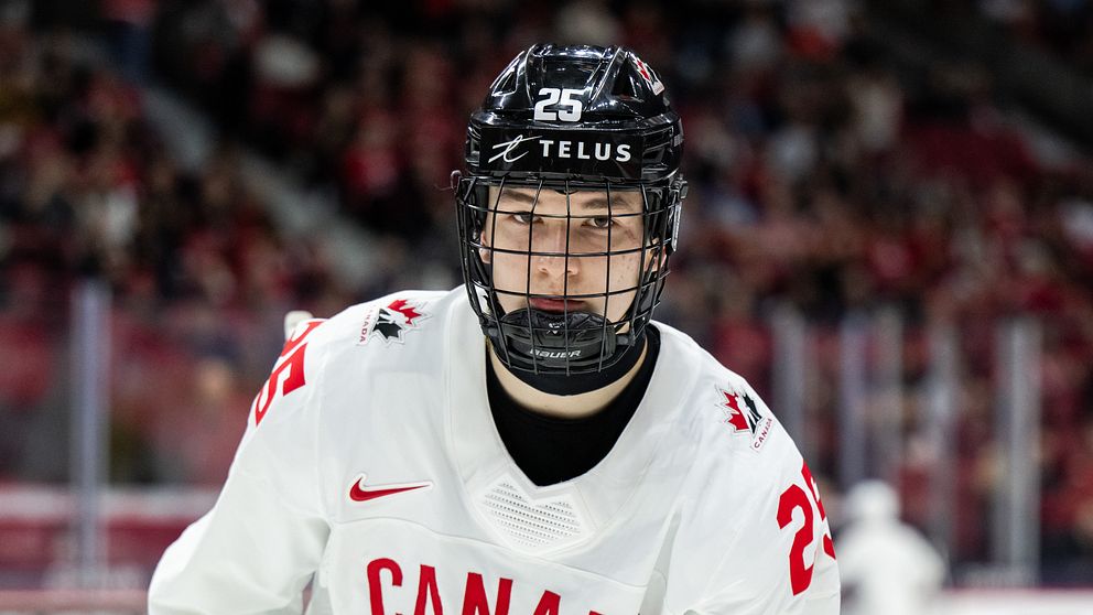 En hockeyspelare i en vit tröja med texten ”CANADA” och nummer 25 på bröstet, står på isen i en arena fylld med åskådare. Spelaren är i en aktiv position, redo att delta i spelet.
