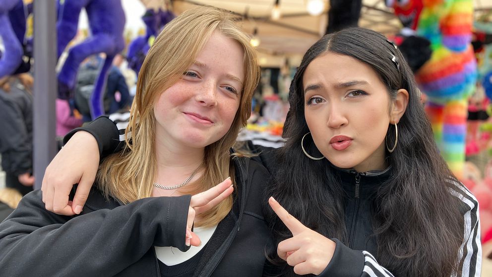 Två personer står tillsammans på en marknad, båda iklädda svarta jackor och poserar med sina armar. I bakgrunden syns färgglada mjukisdjur och en tältstruktur som tyder på en utomhusförsäljning.