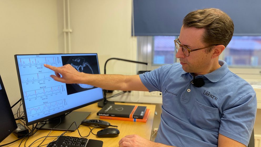 En läkare sitter vid ett skrivbord och pekar mot en datorskärm där en bild på en EKG-kurva syns.