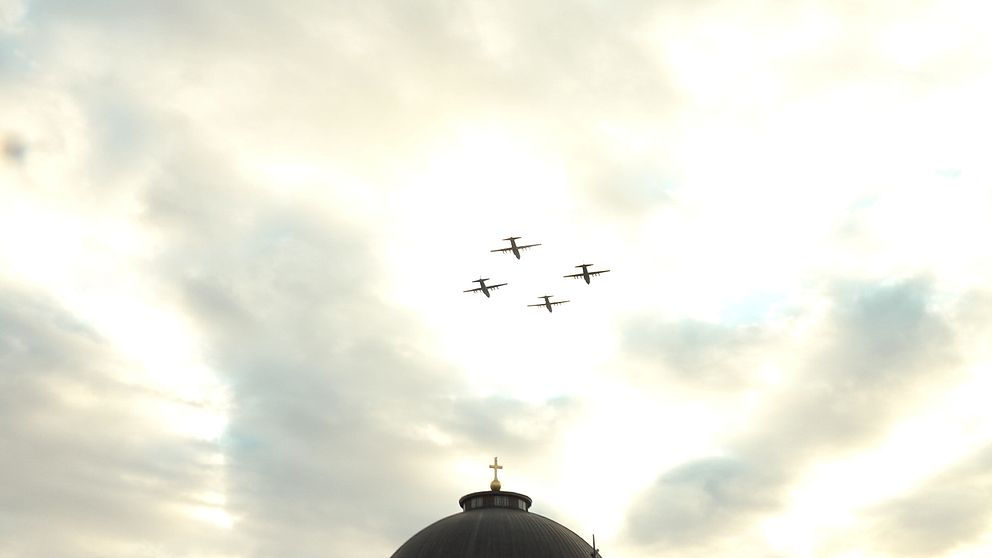 Fyra flygplan passerar över en kupolformad byggnad under en molnig himmel. En gyllene kors syns på toppen av kupolen.