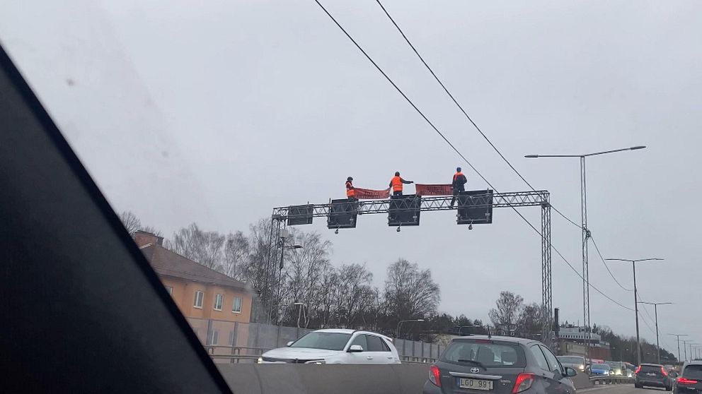 Tre personer med banderoll på stålbalk ovanför motorvägen