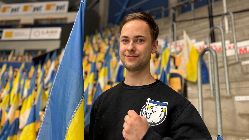 Person i svart tröja med klubbemblem håller blå-gul flagga på läktaren.