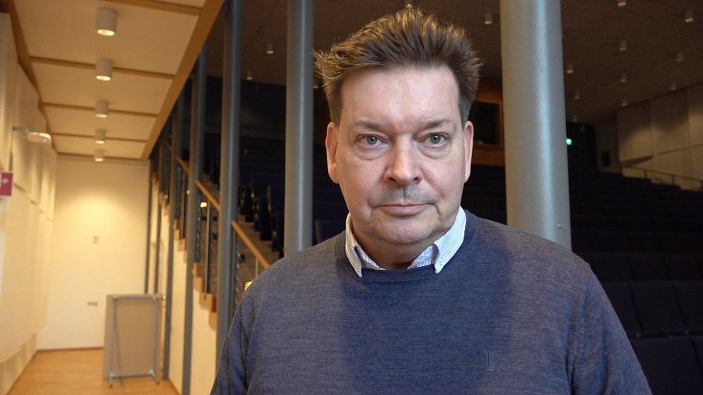 En person iklädd en mörk tröja står i en nästan tom föreläsningssal. I bakgrunden syns en trappa och rader av tomma stolar.