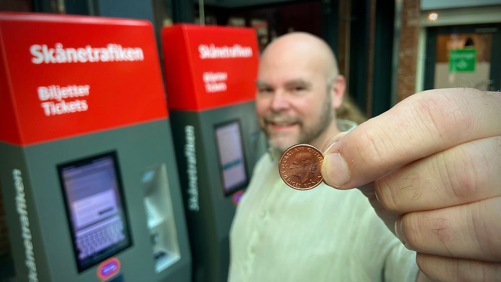 En person håller upp ett mynt framför två biljettautomater från Skånetrafiken. Automaterna har tydlig information om biljettköp på svenska och engelska.