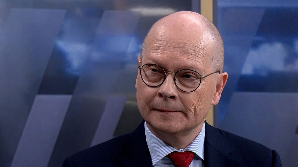 Mats Knutson i kostym med röd slips, bakgrund med suddiga blå och vita mönster.