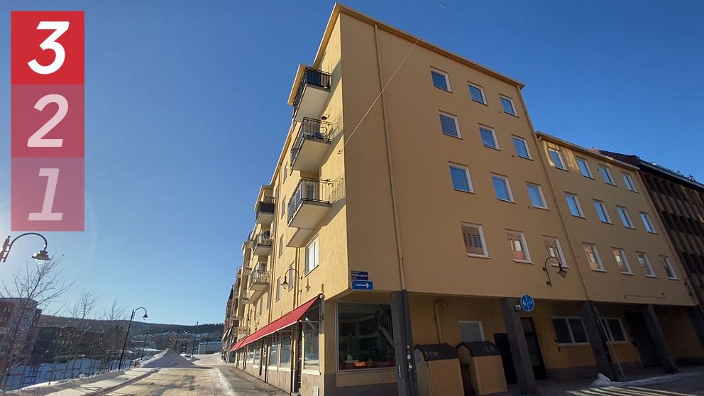 Ett femvåningshus med ljusgul fasad och balkonger ligger på en gata under en klarblå himmel. Gatan är delvis snötäckt och sträcker sig in mot fjärran.