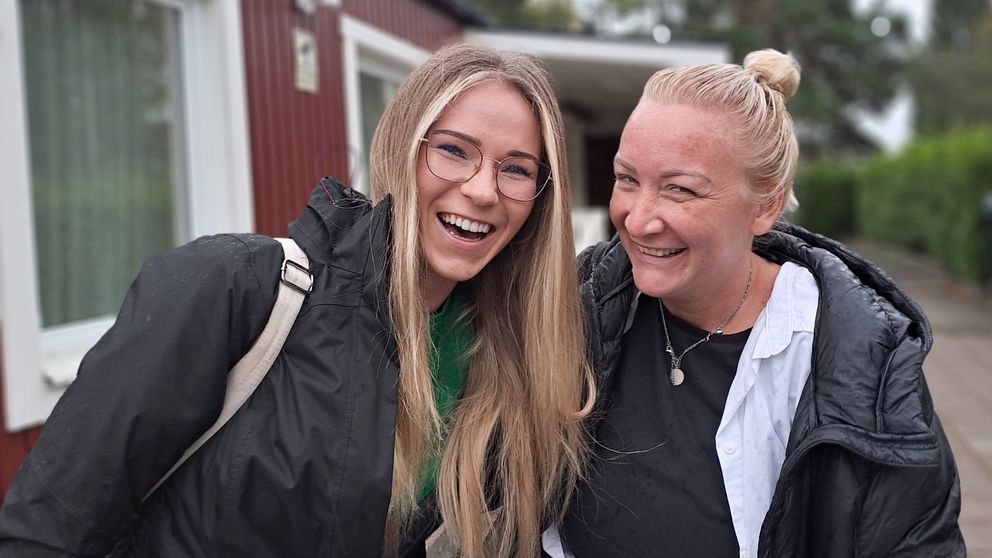 Två kvinnor står arm i arm utanför rött hus i Ronna och skrattar till kameran