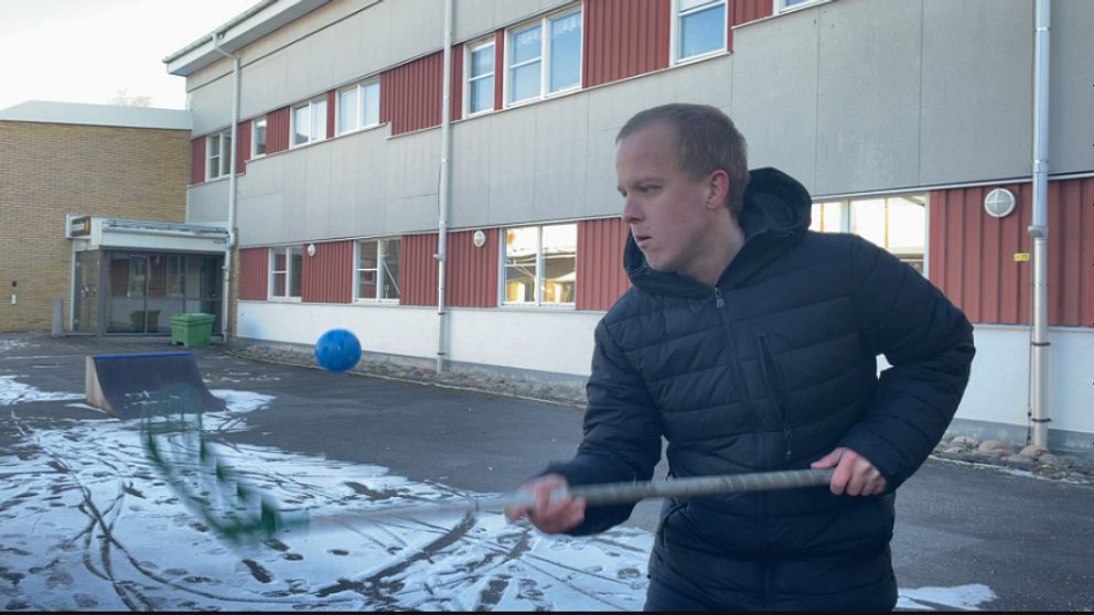 En person som svingar en sportutrustning i en skolgård, med en byggnad i bakgrunden. Snö täcker en del av marken, och flera fönster syns på byggnaden. En blå boll och en ramp för skateboarding eller liknande aktivitet är synliga i scenen.