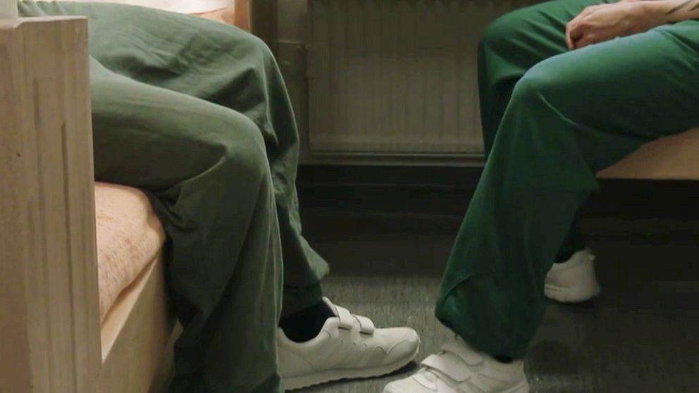 Två personer, anonymiserade, sitter i en häktescell med gröna häkteskläder.