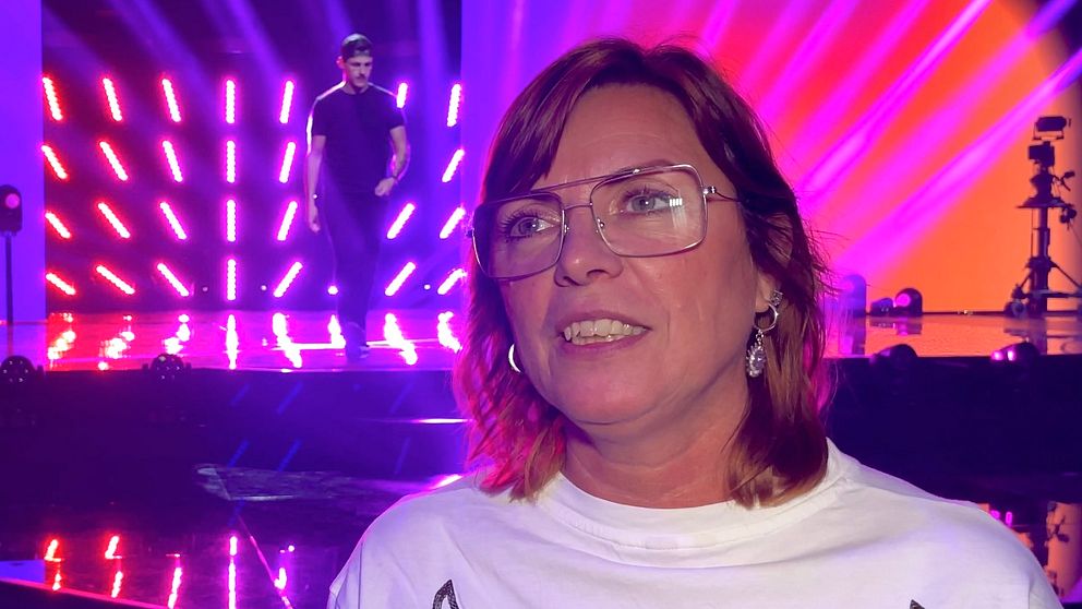 Susanne Bertlin, sångpedagog, står framför en scen med starka ljuseffekter och en manlig artist som uppträder i bakgrunden. Scenen har ljusa, färgglada strålkastare som skapar en livlig atmosfär.