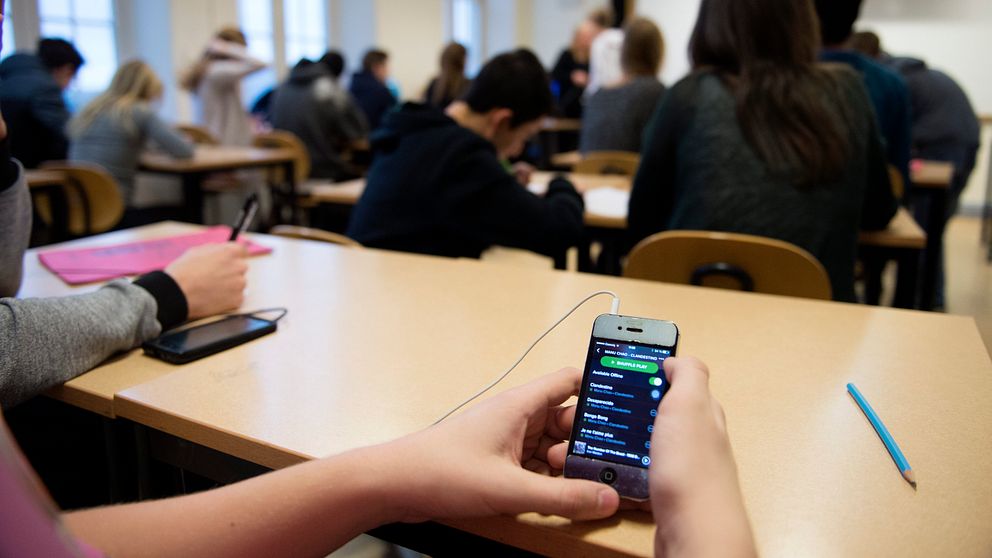 En student sitter på en bänk i ett klassrum och håller en smartphone som visar en musikapp. I bakgrunden syns andra elever som är upptagna med skrivande eller antecknande. Rummet har stora fönster som släpper in naturligt ljus.