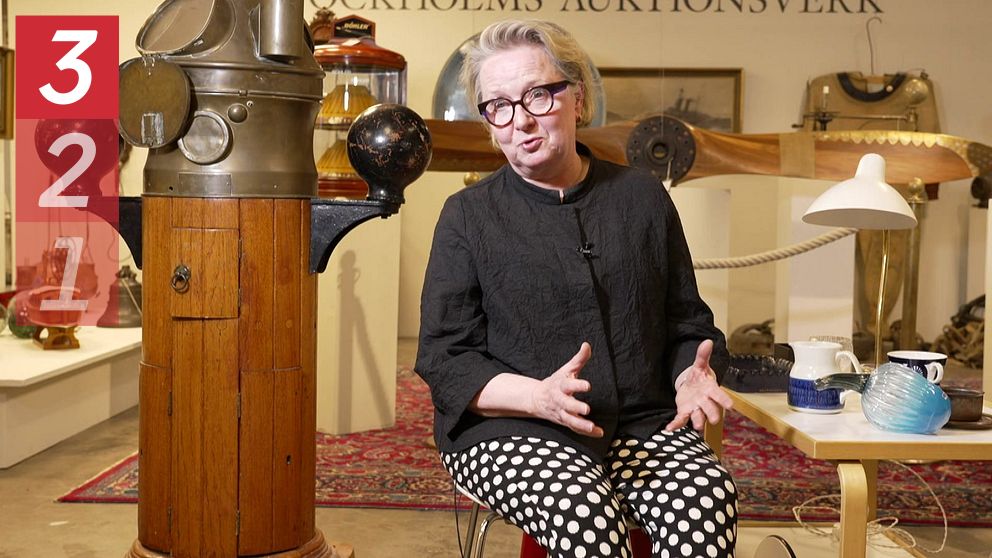 Designexperten Anna-Kaisa Öhgren sitter bredvid maritim utrustning och berättar, bord med porslin i förgrunden.