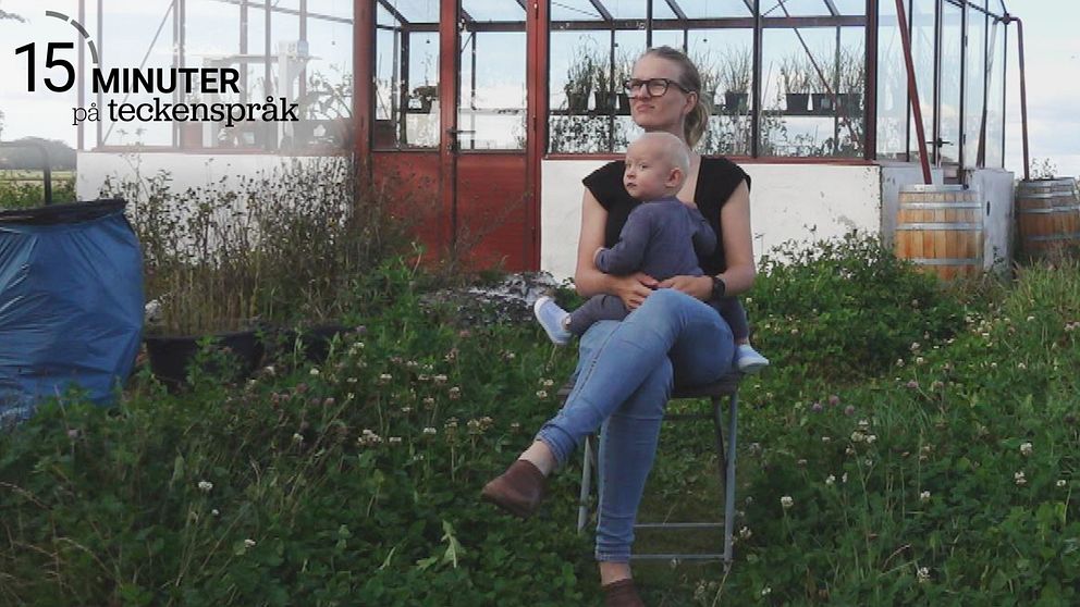 Petra Eklund och hennes cirka 1-åriga son sitter på en stol i en grönskande trädgård framför ett växthus