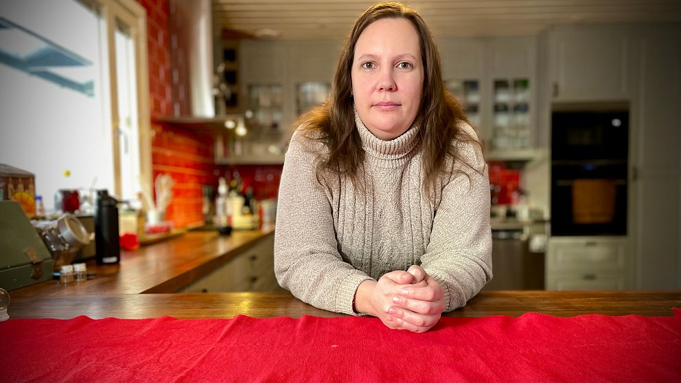 En person i en grå tröja sitter vid ett matbord med en röd duk, omgiven av en hemtrevlig köksmiljö med träelement och bakgrundsdetaljer som köksredskap och hyllor.