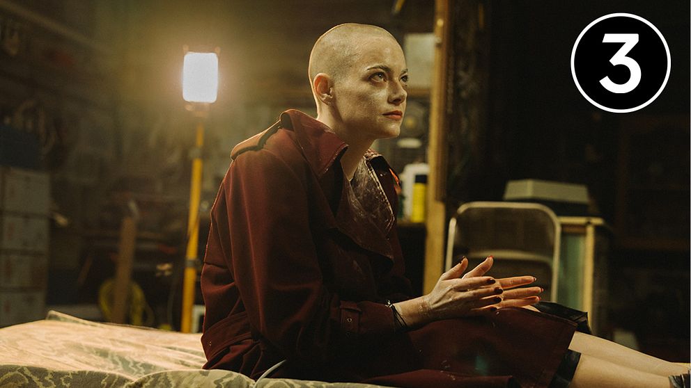 Emma Stone sitter på en säng i en mörk, rörig miljö i filmen Bugonia.